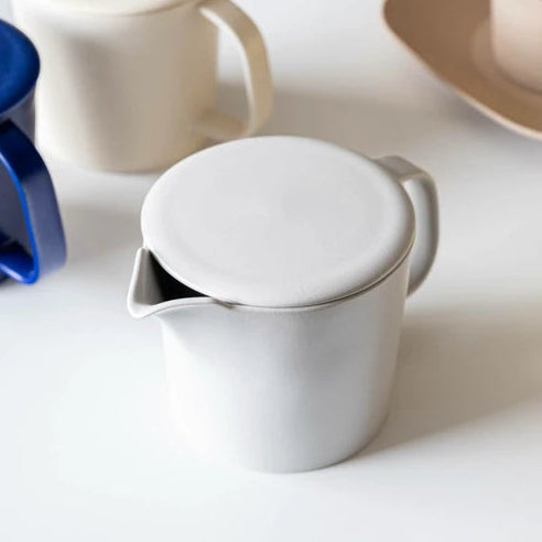 Light grey ceramic teapot with lid on a white surface with other ceramic items in the background.